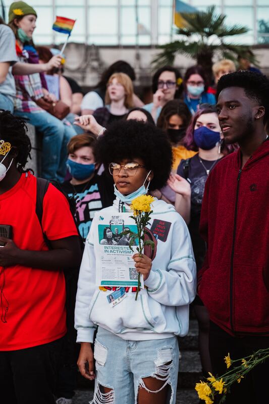 2 Black students stand out at the front of a crowd of young adults. The first student has medium brown skin, a relatively large black afro, glasses, and a light blue mask hanging from her face. In front of her chest, she holds up a BFP flyer and yellow flowers gifted to her from another protester. The second student has dark brown skin and cropped hair and is in a dark red hoodie. He looks off into the distance, his mouth slightly parted as if he was caught in the middle of saying something.